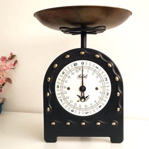 Peut inclure: Une balance de cuisine noire d'époque avec un cadran blanc rond et un bol en bronze. La balance présente un design décoratif avec des clous dorés. Un petit bonsaï aux fleurs roses est à gauche. Mesure en grammes.