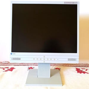 以下が含まれることがあります： 黒い画面の白いコンピューターモニターが、模様入りの布で覆われたテーブルの上にあります。 モニターは銀色のベースと画面の下にボタンが並んでいます。
