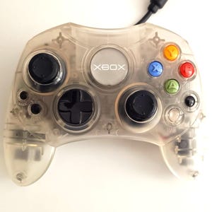 May include: Clear Xbox controller with black buttons, directional pad, and colorful action buttons. The controller has the Xbox logo in the center and a black cable attached. A classic gaming accessory.