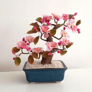 May include: A miniature bonsai tree with pink flowers and brown leaves. The tree is planted in a rectangular blue ceramic pot filled with white pebbles. The bonsai is set against a white background.