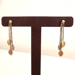 May include: Dangling earrings with gold-colored ball studs and three chains. Each chain has a leaf charm in gold, silver, and rose gold. The earrings are displayed on a brown stand against a white background.