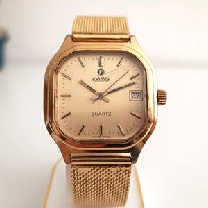Puede incluir: Reloj Roamer Quartz dorado con esfera cuadrada y correa de malla. La esfera del reloj es de color oro claro con manecillas y marcadores negros. La fecha se muestra a las 3 en punto. El reloj está sobre una superficie blanca.