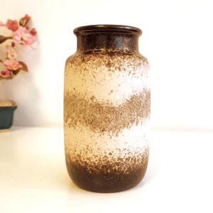 Puede incluir: Un jarrón de cerámica con un diseño texturizado y moteado. El jarrón presenta bandas de colores marrón, blanco y beige. La parte superior e inferior del jarrón son de color marrón oscuro. El jarrón es cilíndrico con un borde ligeramente ensanchado.