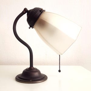 Może przedstawiać: Lampa biurkowa w stylu antycznym z brązowym wykończeniem. Lampa posiada zakrzywione ramię i klosz z matowego szkła. Łańcuszek zwisa z klosza. Lampa stoi na białej powierzchni.