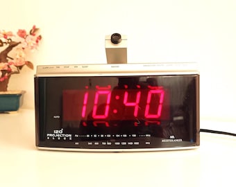 May include: A digital alarm clock displaying 10:40 in red numerals. The clock has a silver frame and a black face. A small bonsai tree with pink flowers sits to the left. The clock has a projection feature.