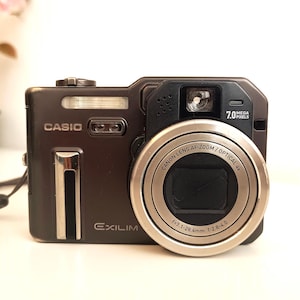Könnte beinhalten: Dunkelbraune Casio Exilim Digitalkamera mit silbernem Objektiv und Blitz. Die Kamera hat eine Handschlaufe und das Objektiv trägt die Aufschrift "Canon Lens AF Zoom / Optical 4x". Die Kamera hat 7,0 Megapixel.