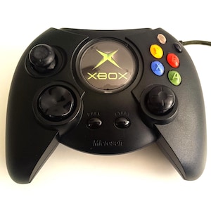 May include: Black Xbox controller featuring the Xbox logo. It has two analogue sticks, a directional pad, and coloured buttons. The word "Microsoft" is printed on the bottom of the controller.
