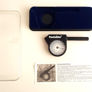 May include: A vintage Panchelidon instrument set, comprising a black and silver compass, a dark blue case, a clear plastic case, and a printed instruction sheet. The compass has a circular dial and a handle.