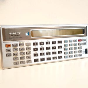 Könnte beinhalten: Silberner wissenschaftlicher Taschenrechner Sharp FL-5100 mit einem Display und zahlreichen Tasten. Der Rechner hat ein silbernes Gehäuse mit schwarzen und weißen Tasten, einschließlich Zahlen, Funktionen und wissenschaftlichen Notationen. Der Rechner steht auf einer weißen Oberfläche.