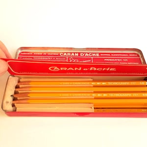 Puede incluir: Un estuche de lápices de metal rojo con el nombre de la marca "Caran d'Ache" en letras blancas. El estuche está abierto, revelando varios lápices amarillos con el texto "Technograph 777 HB".