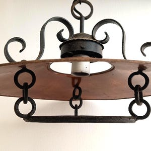 May include: An antique hanging light fixture with a copper-colored shade and black wrought iron accents. The fixture has a circular shade with a central opening for the light bulb and is suspended by chains.