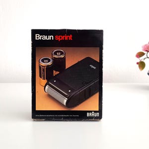 以下が含まれることがあります： ヴィンテージのブラウン・スプリント電気シェーバーのオリジナルボックス。黒い箱には、ブランド名と製品名が白と赤で表示されています。箱の画像には、シェーバーと2つのバッテリーが写っています。背景には、ピンクの花が咲く小さな盆栽があります。