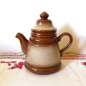 May include: A brown ceramic teapot with a lid and handle. The teapot has a rounded body and a spout. The color gradient goes from light beige to dark brown. The teapot is sitting on a white tablecloth with red floral embroidery.