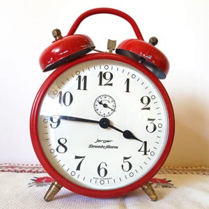 May include: A vintage red Jerger Drumbealarm clock with a white face and black numerals. The clock has a round shape with a red frame, a curved handle, and two bells on top. The clock's hands point to 4:19.