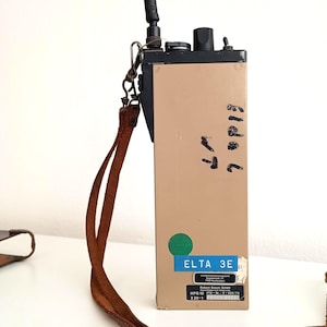 Op de afbeelding: Een beige ELTA 3E radio met een zwarte bovenkant, antenne en leren riem. De radio heeft een groene cirkel en een blauw label met tekst. Handgeschreven nummers staan op de zijkant.