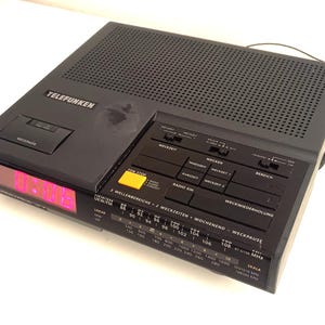 Könnte beinhalten: Ein schwarzer digitaler Telefunken-Wecker mit roter Digitalanzeige. Der Wecker verfügt über mehrere Tasten und Drehregler, darunter eine Schlummerfunktion und Radiosteuerung. Die Oberseite hat ein perforiertes Lautsprechergitter.