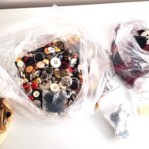 Puede incluir: Una colección de botones variados en una bolsa de plástico transparente, incluyendo botones blancos, negros, rojos y marrones. También se ven otras bolsas de botones, incluyendo una bolsa de botones rojos, y un paquete de horquillas doradas.
