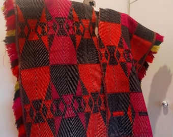 Könnte beinhalten: Ein rotes, schwarzes und pinkfarbenes gewebtes Poncho mit Fransen. Das Poncho hat ein geometrisches Muster und wird auf einem Holzständer präsentiert.