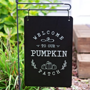 Puede incluir: Un letrero de jardín rectangular negro con las palabras "WELCOME TO OUR PUMPKIN PATCH" en escritura blanca. El letrero está colgado de una estaca de metal negro, con un remate decorativo en la parte superior. El letrero se exhibe al aire libre, rodeado de vegetación.