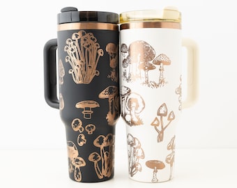 May include: Two stainless steel travel mugs with a mushroom pattern. The mug on the left is black with a copper-coloured mushroom design. The mug on the right is white with a copper-coloured mushroom design.