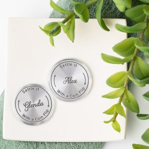 May include: Two silver decision coins with the text "Settle It" and "With a Coin Flip" around the edges. One coin has "Glenda" and the other has "Alex" in the center. The coins are on a white surface with green foliage.