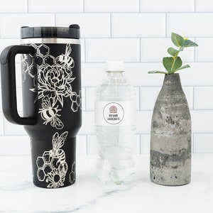 Stanley brand 40 oz tumbler in black with bumble bee and honeycomb design wrapped around the tumbler. Shown with a water bottle for size comparison.