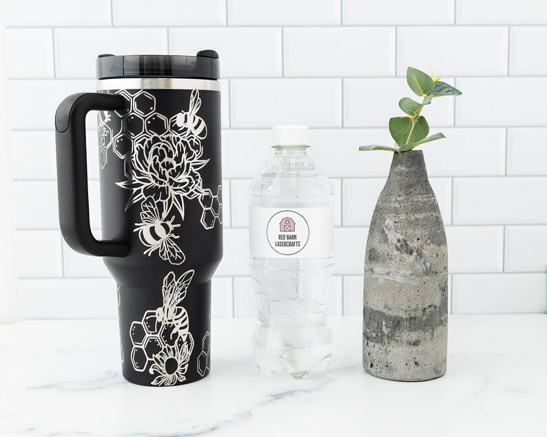 Stanley brand 40 oz tumbler in black with bumble bee and honeycomb design wrapped around the tumbler. Shown with a water bottle for size comparison.