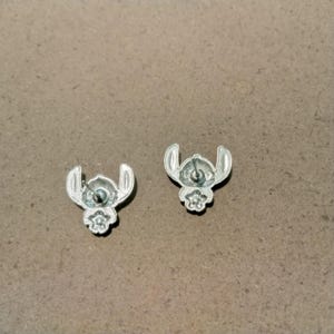 May include: A pair of silver-colored stud earrings. Each earring features a stylized design of a character with large ears and a small flower-like shape at the bottom. The earrings are set against a neutral background.