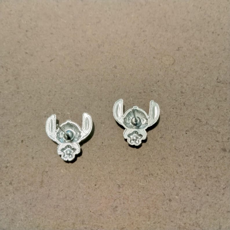 May include: A pair of silver-colored stud earrings. Each earring features a stylized design of a character with large ears and a small flower-like shape at the bottom. The earrings are set against a neutral background.