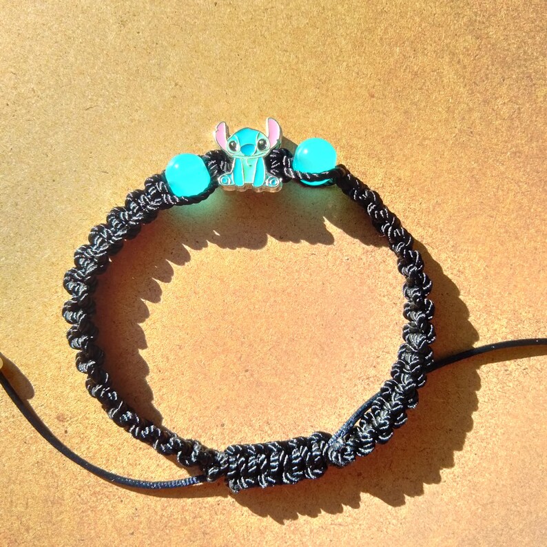 May include: Black macrame bracelet featuring a turquoise Stitch charm and two turquoise beads. The charm depicts the character Stitch from the animated film Lilo & Stitch. The bracelet is knotted with a sliding knot closure.