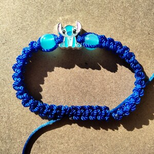 May include: A blue macrame bracelet featuring a Stitch charm and two blue beads. The bracelet is made of woven blue cord and has a sliding knot closure. The Stitch charm is a light blue and silver metal.