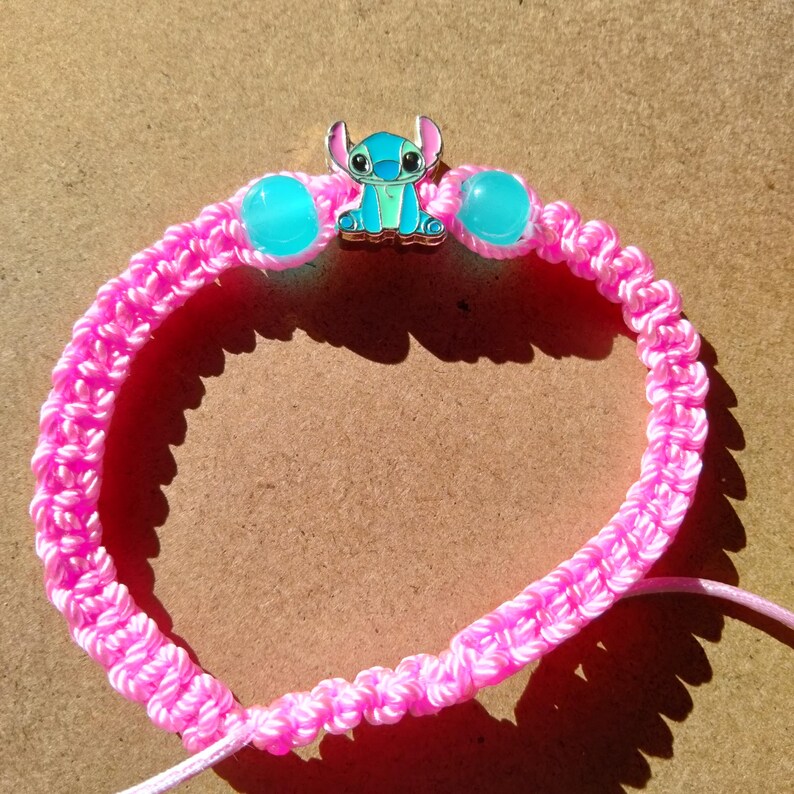 May include: A pink macrame bracelet featuring a blue and silver Stitch charm and two turquoise beads. The bracelet is made of woven pink cord and has a sliding knot closure. The charm depicts the character Stitch from the Disney movie Lilo & Stitch.