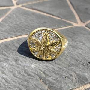 Puede incluir: Un anillo de oro con un gran diseño de hoja de marihuana incrustado con diamantes de imitación transparentes.