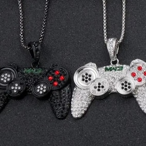 May include: Two pendant necklaces featuring a video game controller design. One is black with red and white accents, the other is silver with red and black accents. Both have a chain and are encrusted with rhinestones.