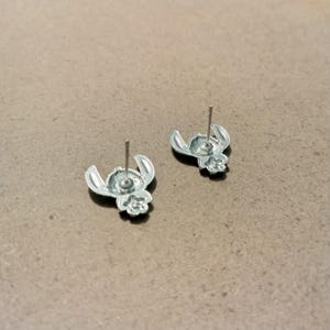 May include: Two silver-colored stud earrings featuring a cartoon character design. The earrings are in the shape of a character's head with large ears and a small nose. The posts are silver and extend from the back of the earrings.