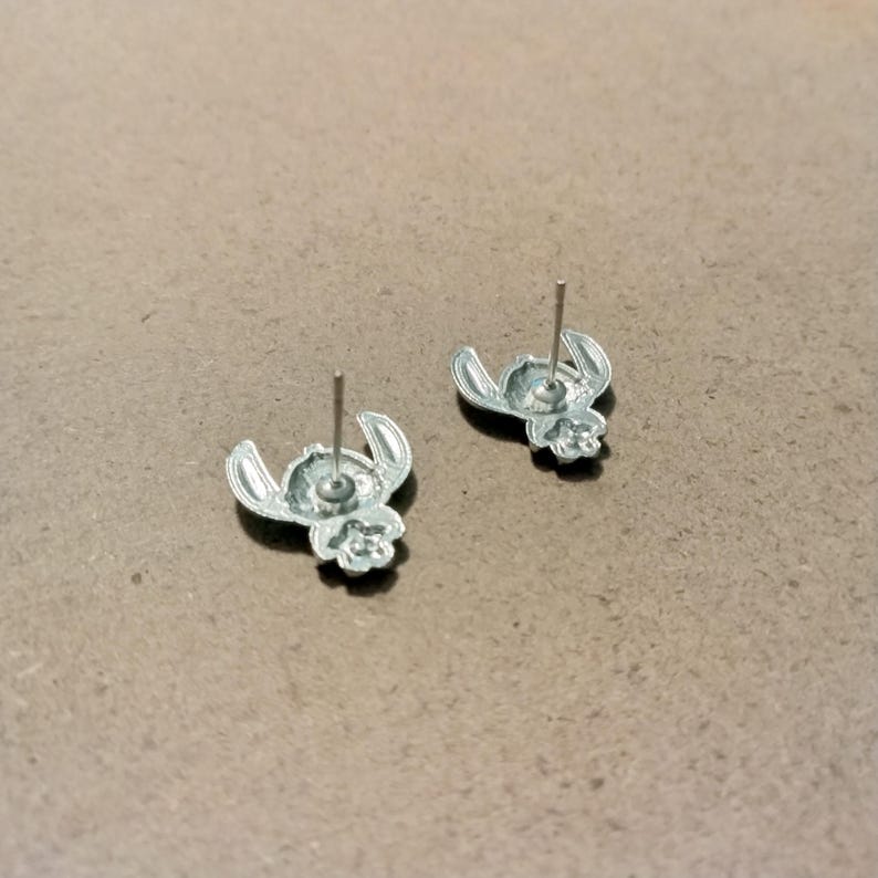 May include: Two silver-colored stud earrings featuring a cartoon character design. The earrings are in the shape of a character's head with large ears and a small nose. The posts are silver and extend from the back of the earrings.