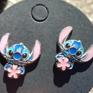 May include: A pair of blue and pink enamel stud earrings featuring the character Stitch from Lilo & Stitch. The earrings have a silver-colored outline and a pink flower detail.
