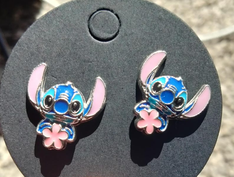 May include: A pair of blue and pink enamel stud earrings featuring the character Stitch from Lilo & Stitch. The earrings have a silver-colored outline and a pink flower detail.