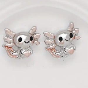 Puede incluir: Un par de pendientes de plata con forma de ajolote con detalles en rosa. Los ajolotes tienen ojos negros y una expresión sonriente.