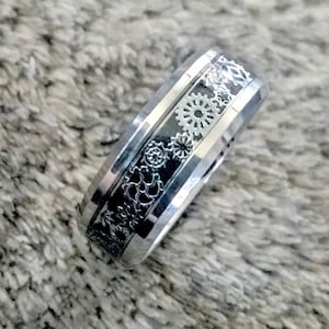 May include: A silver ring with a black band and a design of gears and other mechanical elements.