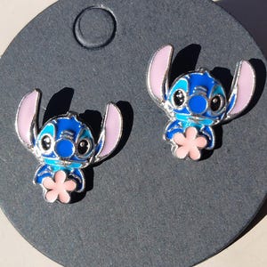 May include: A pair of blue and pink enamel Stitch stud earrings. The earrings feature a silver outline and a pink flower. The earrings are on a dark gray background.