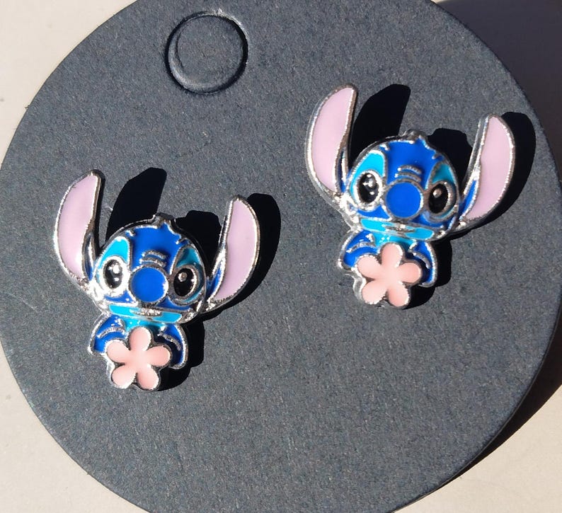 May include: A pair of blue and pink enamel Stitch stud earrings. The earrings feature a silver outline and a pink flower. The earrings are on a dark gray background.