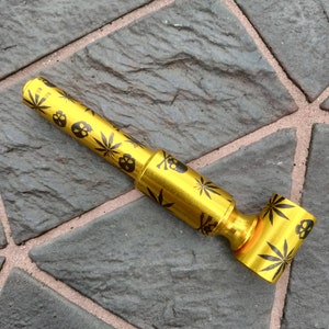 May include: A gold-coloured metal pipe with a skull and marijuana leaf design. The pipe is on a grey stone surface.