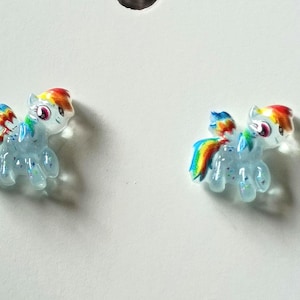 May include: A pair of stud earrings shaped like small ponies with translucent light blue bodies, glitter accents, and vibrant rainbow-colored manes and tails. The ponies feature large eyes and are depicted in a standing position with wings.