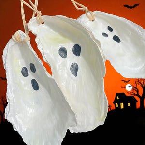 May include: Three white ghost decorations with black eyes and mouths hang against an orange background. The Halloween-themed decorations are made from oyster shells and are suspended by natural twine. A spooky scene with a house, trees, bats, and a moon is in the background.