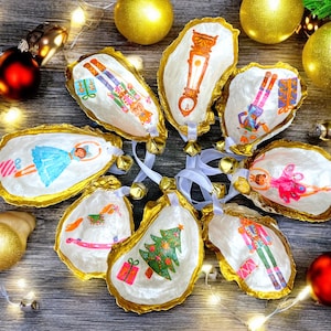 May include: A collection of handmade Christmas ornaments crafted from oyster shells. Each shell features a unique hand-painted design, including a ballerina, a clock, and a Christmas tree. The ornaments are trimmed with gold and adorned with small bells and ribbons.