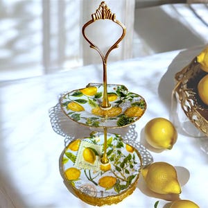 May include: Two-tiered cake stand with a gold metal frame and a white base with a lemon print. The stand is decorated with gold accents and features a delicate lace design. The stand is filled with lemons.