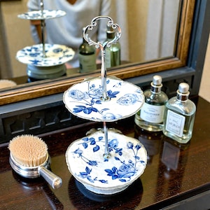 May include: A two-tiered serving tray with a silver handle and floral design. The white ceramic plates feature blue rose patterns. The tray is displayed with a silver-handled brush and two glass perfume bottles.