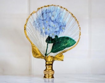 May include: A decorative lamp finial featuring a seashell design with a painted blue hydrangea flower and green leaves. The finial has a gold-colored base and accents, adding a touch of elegance to any lamp.