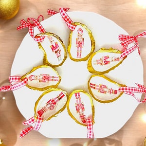 May include: Christmas ornaments made from oyster shells, featuring nutcracker designs. The shells have gold-painted edges and are adorned with red and white checkered ribbons for hanging. Gold Christmas balls and string lights are in the background.